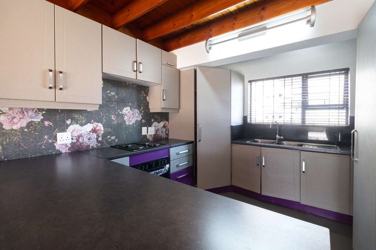 A functional kitchen featuring dark countertops, light-toned cabinetry with purple accents, floral tile backsplash, and exposed timber ceiling beams.