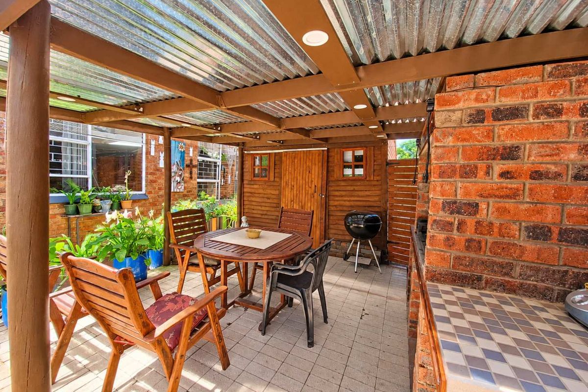 A covered outdoor patio area featuring a built-in brick fireplace, tiled prep counter, and corrugated roofing, providing a functional space for outdoor dining and entertaining.