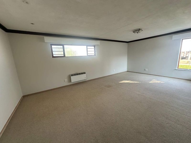 A spacious, empty living area featuring neutral walls, carpeted flooring, and a wall-mounted electric heater, offering a blank canvas for interior design.