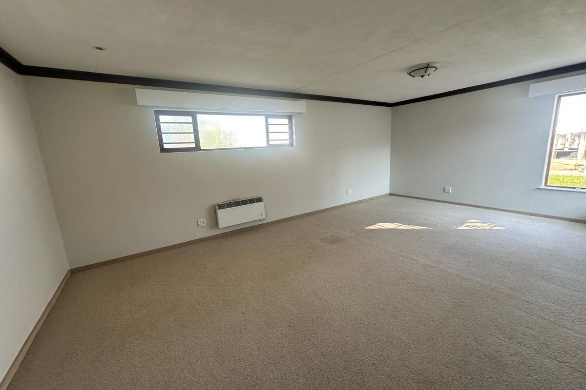 A spacious, empty living area featuring neutral walls, carpeted flooring, and a wall-mounted electric heater, offering a blank canvas for interior design.