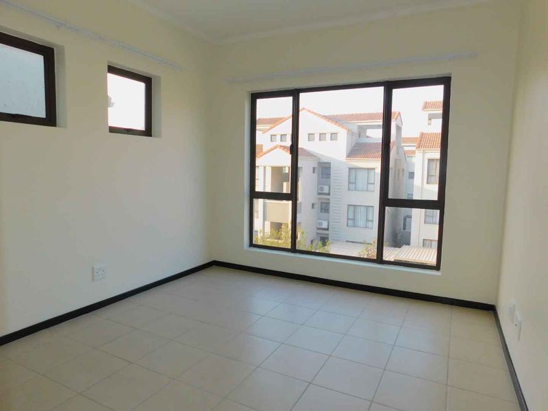 A bright, empty bedroom featuring neutral tiled flooring and large aluminium-framed windows providing natural light and a view of the surrounding complex.