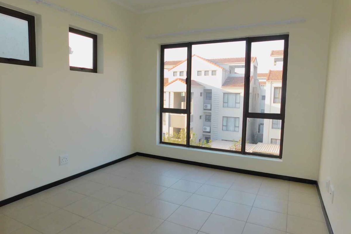 A bright, empty bedroom featuring neutral tiled flooring and large aluminium-framed windows providing natural light and a view of the surrounding complex.