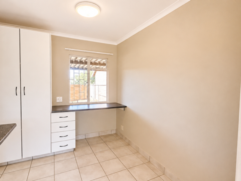 A clean, functional scullery or utility area featuring built-in white cabinetry, a dark laminate workspace, tiled flooring, and a window with security bars.