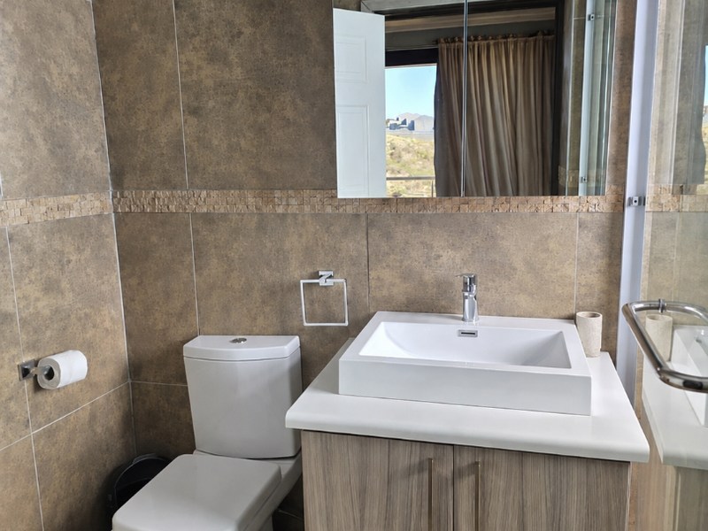 A modern bathroom featuring a vessel sink vanity, wall-mounted mirror cabinet, toilet, and a glass-enclosed shower with neutral earth-toned wall and floor tiles.