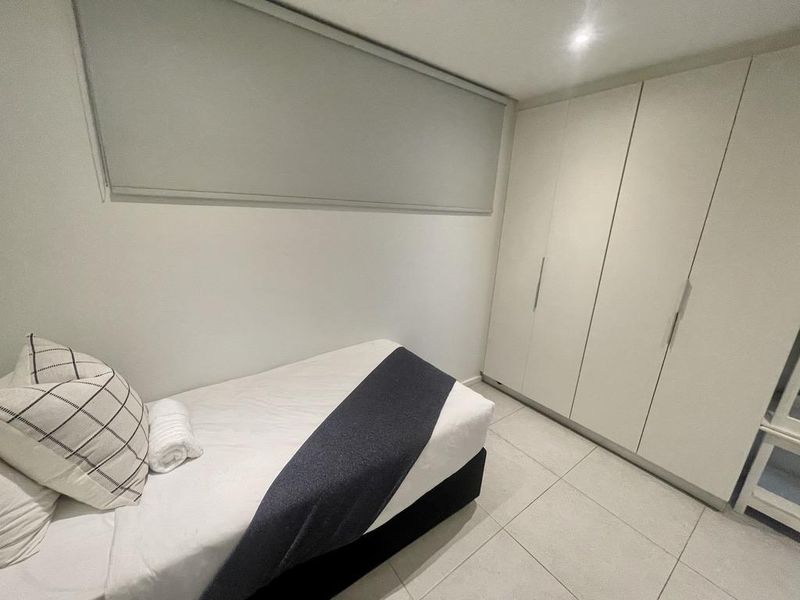 A clean and modern bedroom featuring built-in floor-to-ceiling wardrobes, neutral tiled flooring, and a roller blind for privacy.