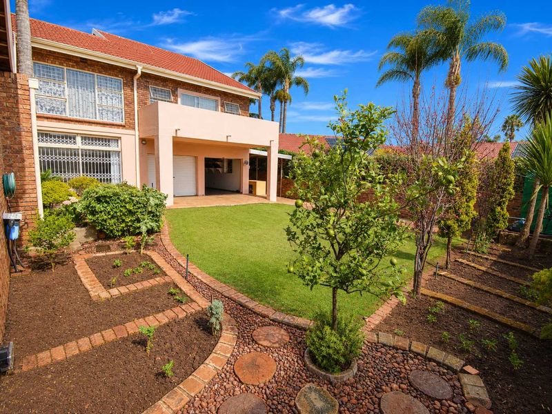 A well-maintained rear garden featuring a manicured lawn, landscaped brick-edged garden beds, and a covered patio area attached to the two-story brick residence.