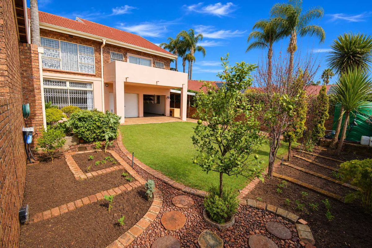 A well-maintained rear garden featuring a manicured lawn, landscaped brick-edged garden beds, and a covered patio area attached to the two-story brick residence.