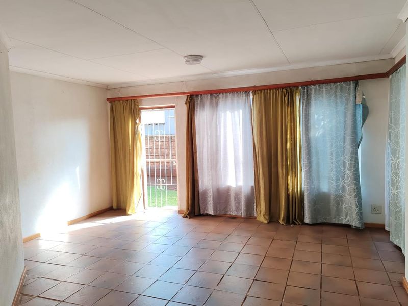 An empty living room featuring tiled flooring, large windows with curtain rails, and a security gate providing access to the exterior.