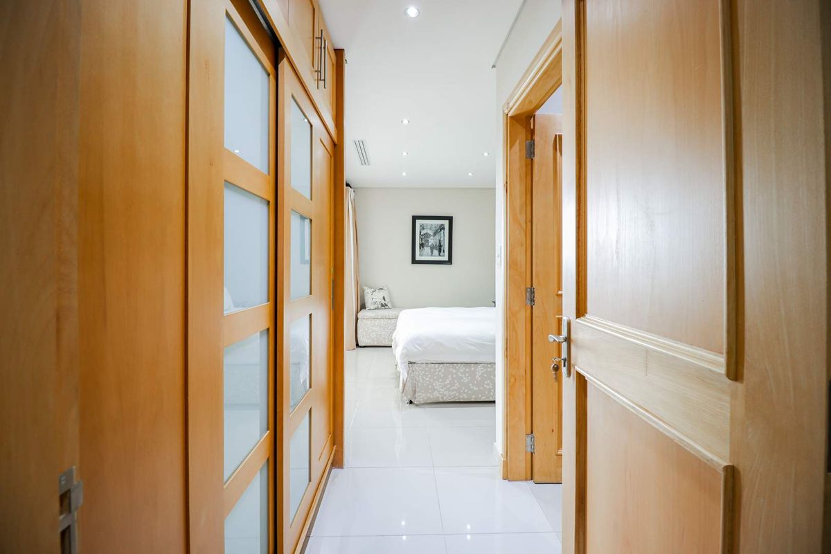 A well-maintained bedroom featuring built-in wooden cupboards, polished tiled flooring, and recessed ceiling lighting, viewed from the hallway.