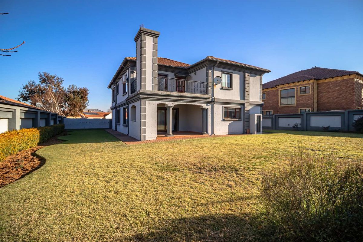 Rear view of a two-story house featuring a covered patio, upper-level balcony, chimney, and a spacious lawn area enclosed by a perimeter wall.