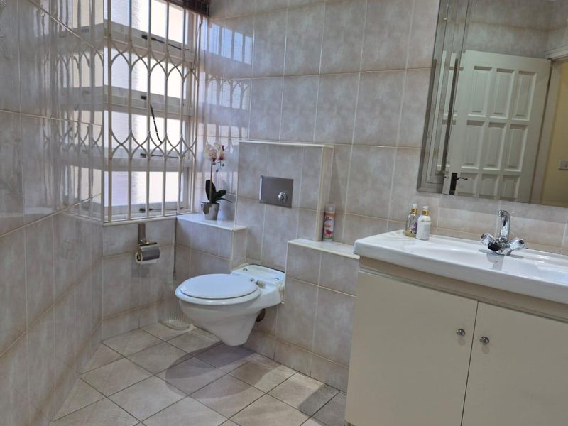 A functional bathroom featuring a wall-mounted toilet, vanity cabinet with basin, and tiled finishes throughout, complemented by a window with security bars.