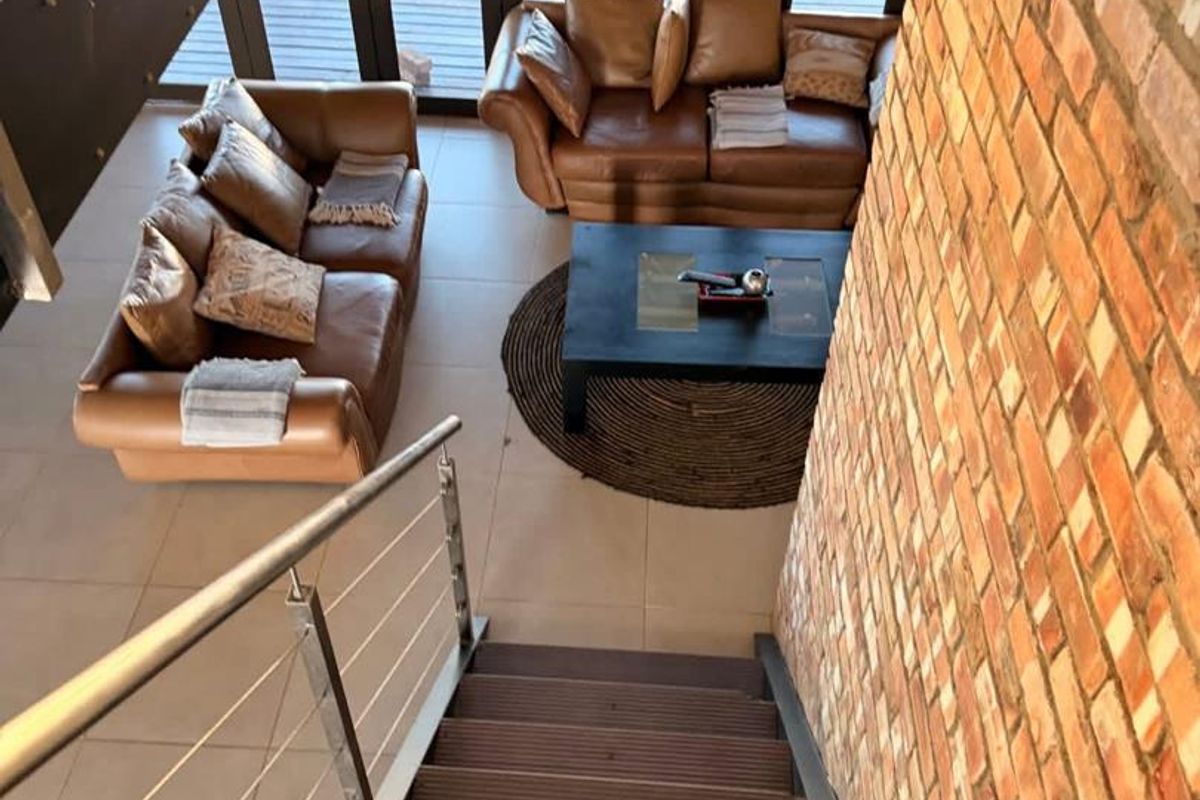 A view from the staircase looking down into a spacious living area featuring exposed brick walls, tiled flooring, and large glass doors leading outside.