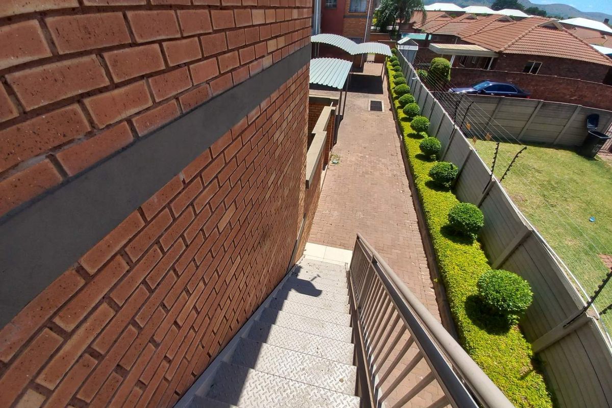 An elevated view looking down a metal staircase towards a paved side walkway, featuring brick exterior walls and a manicured hedge boundary.