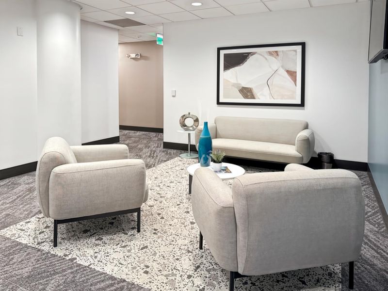 A professional office waiting or breakout area featuring a modern seating arrangement, neutral color palette, and a combination of carpet and terrazzo-style flooring.