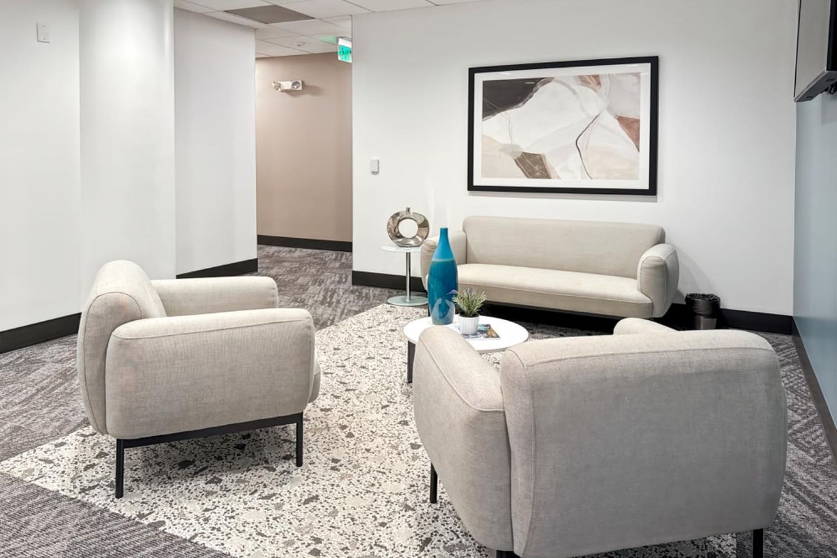 A professional office waiting or breakout area featuring a modern seating arrangement, neutral color palette, and a combination of carpet and terrazzo-style flooring.