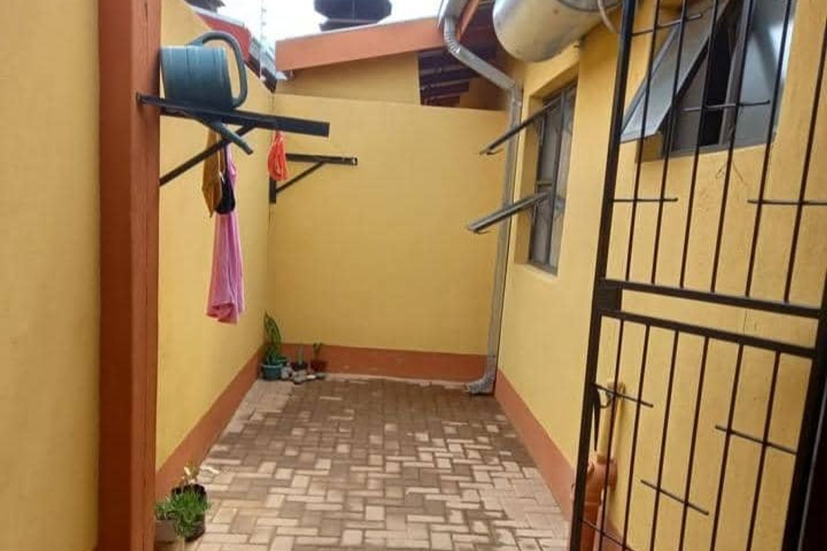 A paved exterior utility area featuring a wall-mounted drying rack, external geyser, and security gate access.