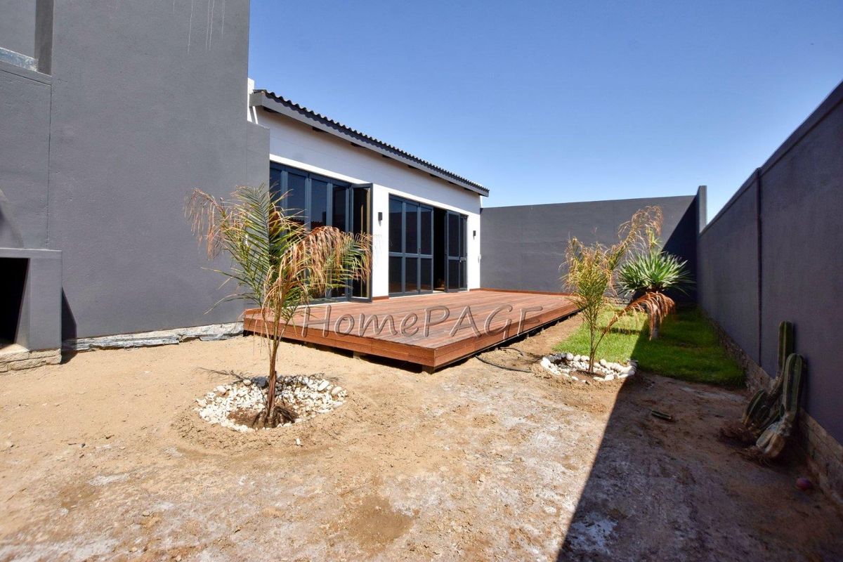 A rear exterior view featuring a wooden deck, dark-framed sliding glass doors, and a private courtyard with perimeter walls.