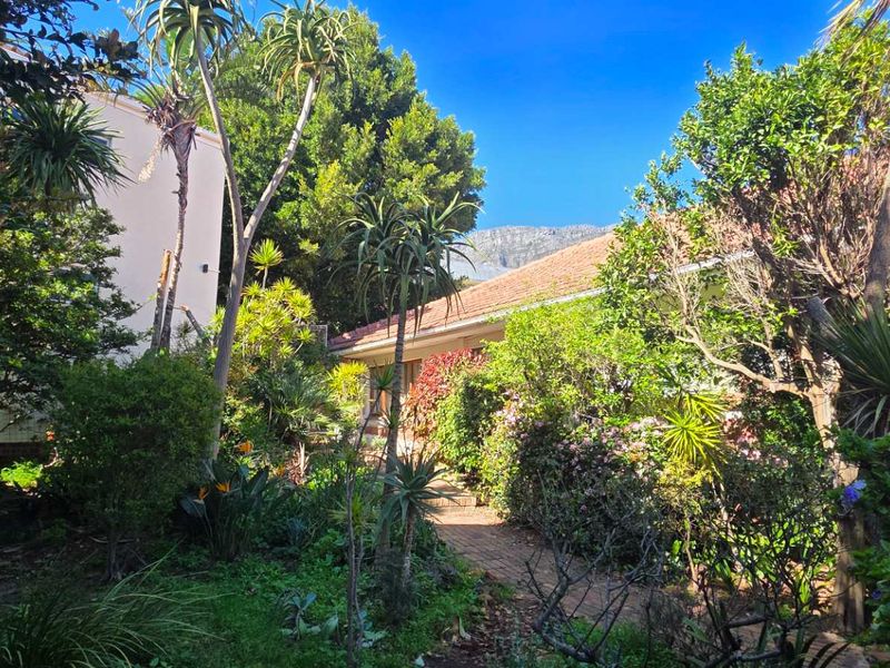 A lush, established garden featuring a brick pathway leading towards the house, surrounded by mature trees and shrubs.