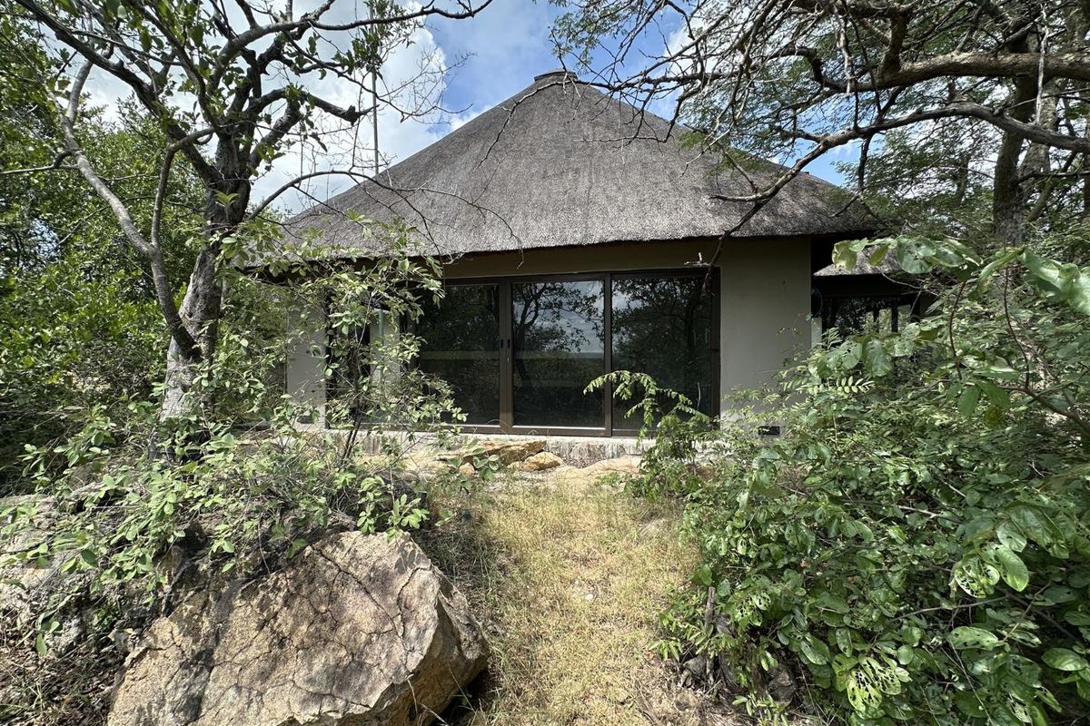 A detached dwelling featuring a traditional thatched roof and large sliding glass doors, nestled within a natural, bushveld-style landscape.