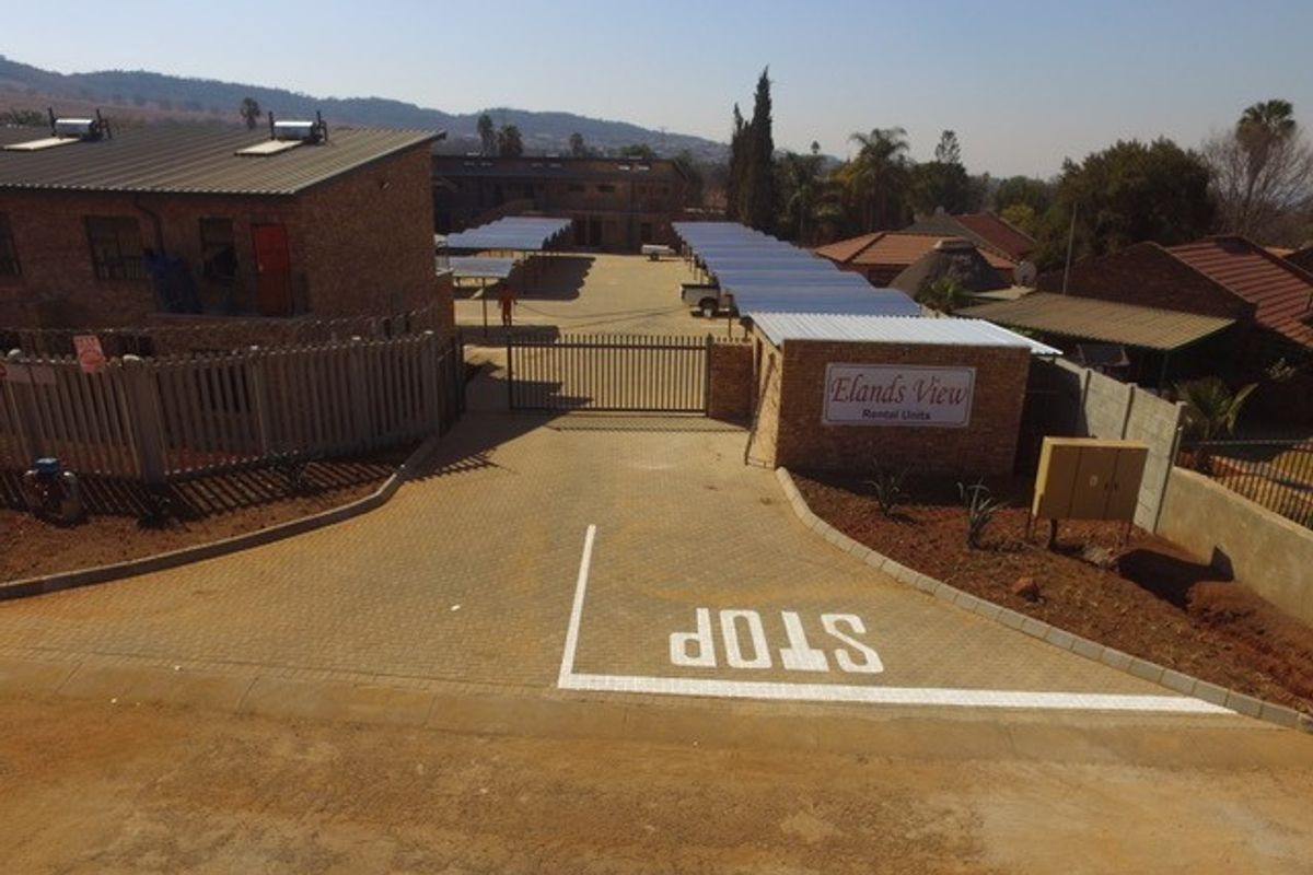 Aerial view of the apartment complex entrance, featuring a paved driveway, security gate, and covered parking bays.