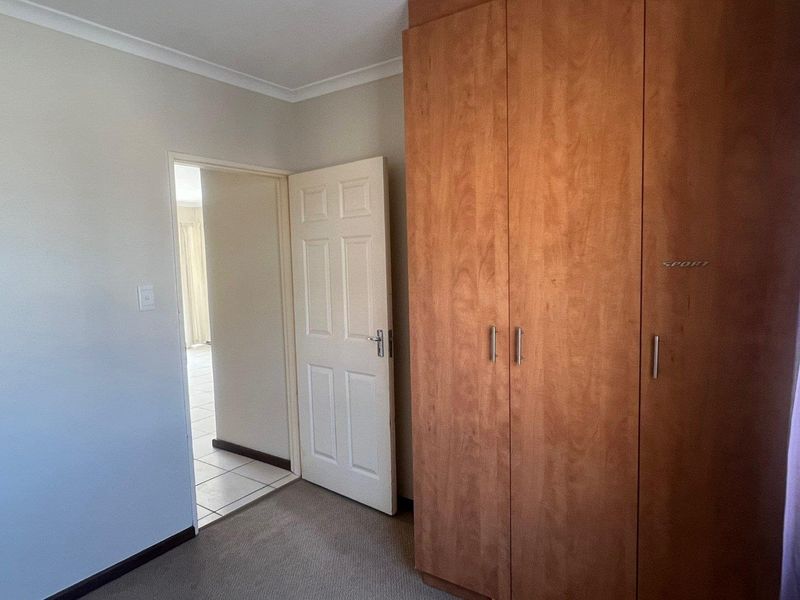 A bedroom featuring built-in wooden cupboards, neutral wall finishes, and carpeted flooring, with a doorway leading to an adjacent tiled living area.