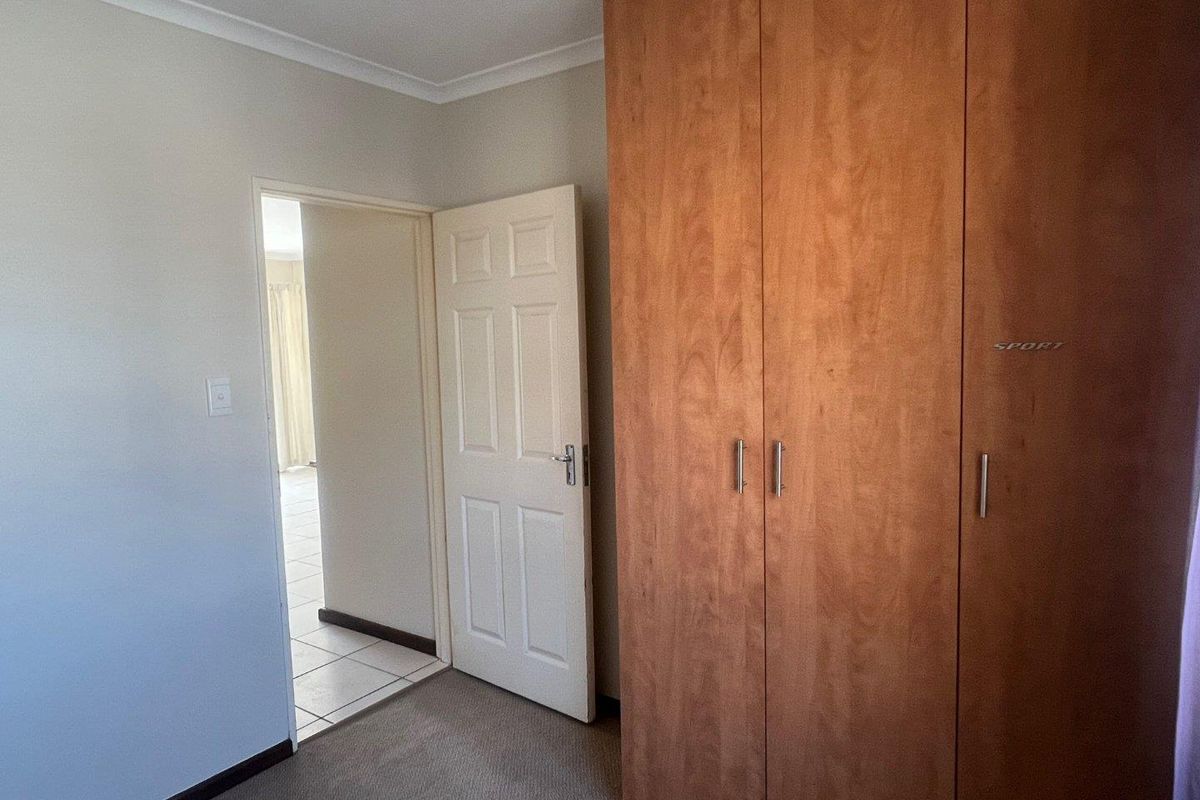 A bedroom featuring built-in wooden cupboards, neutral wall finishes, and carpeted flooring, with a doorway leading to an adjacent tiled living area.