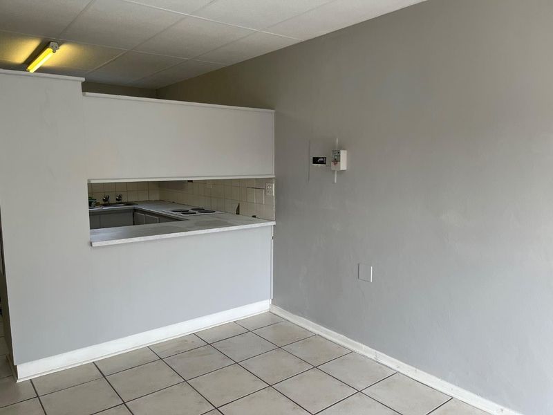 An empty open-plan living area featuring tiled flooring, a drop ceiling, and a functional kitchen pass-through with an integrated electric hob.