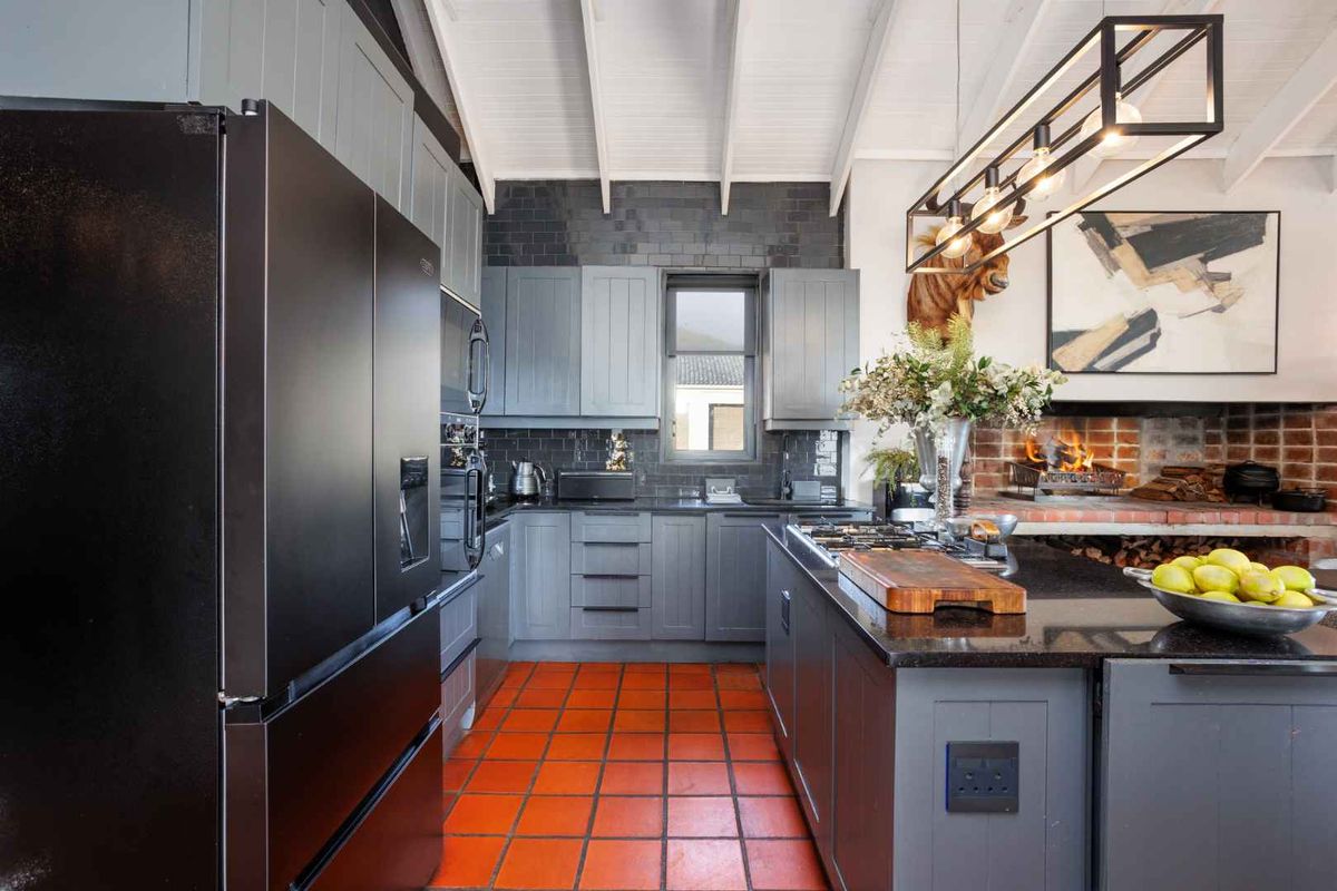 Modern farmhouse kitchen featuring dark grey cabinetry, granite countertops, a gas hob, and a rustic brick fireplace under an exposed beam ceiling.