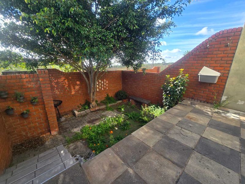 A private rear courtyard featuring a paved patio area, brick boundary walls, and a small established garden with a mature tree.