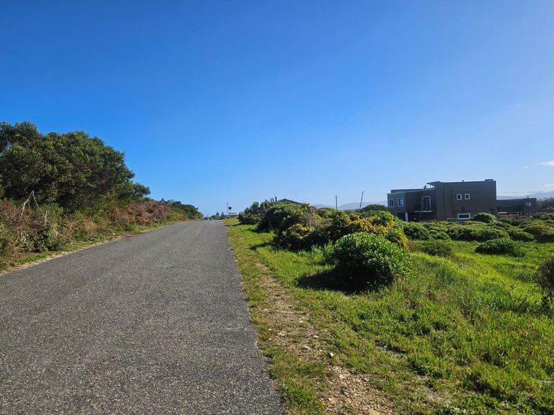 A view of the asphalt access road leading towards the property, surrounded by natural coastal vegetation and open land.