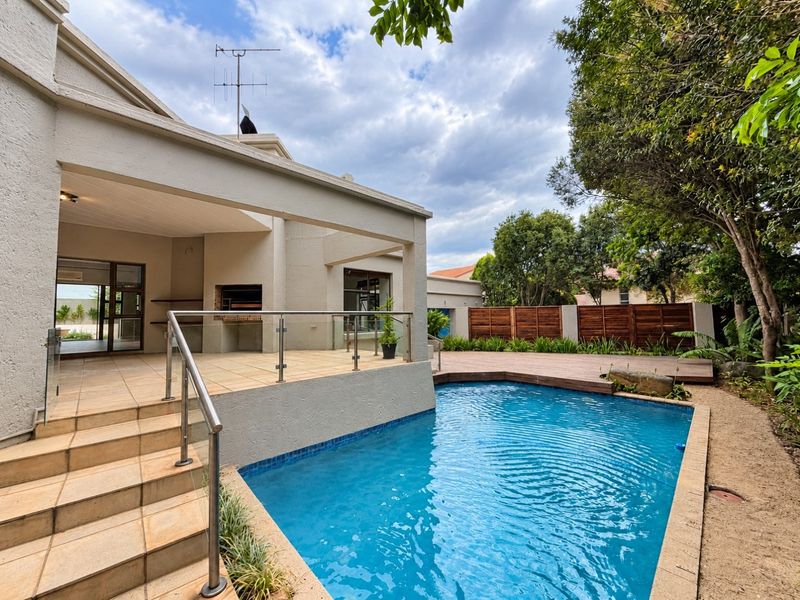 Outdoor entertainment area featuring a swimming pool, wooden decking, and a covered patio with a built-in braai and glass balustrade.