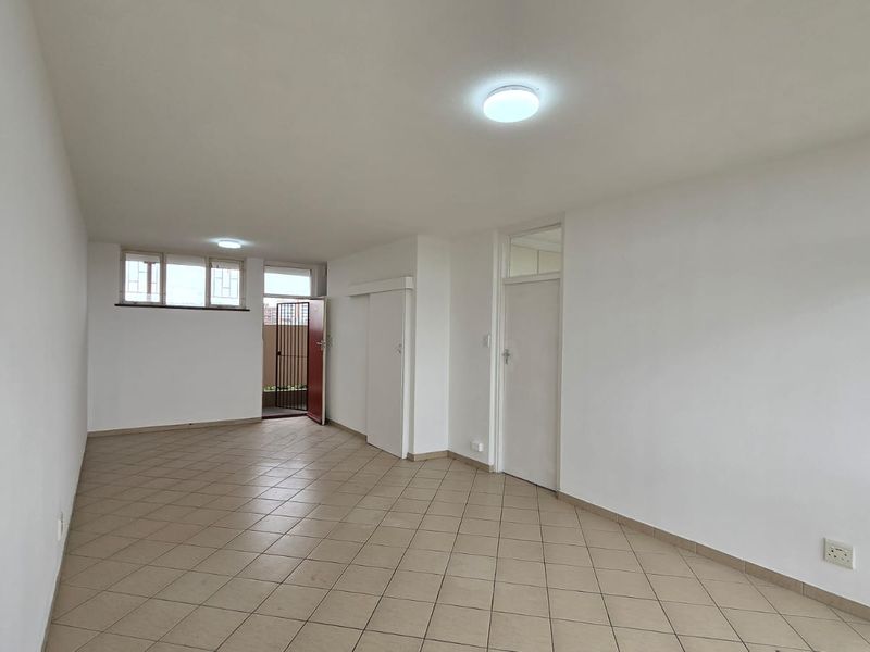 A bright, empty living area featuring neutral ceramic floor tiles, clean white walls, and direct access to an outdoor space through a security gate.