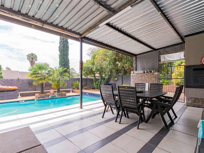 Covered patio area featuring tiled flooring and a built-in braai, overlooking a private swimming pool and landscaped garden.