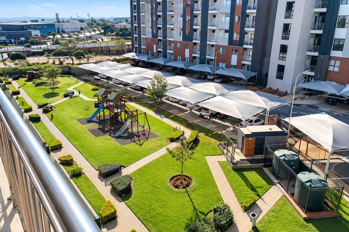 Aerial view of the apartment complex grounds featuring landscaped gardens, a children's playground, and covered parking bays with shade-cloth structures.