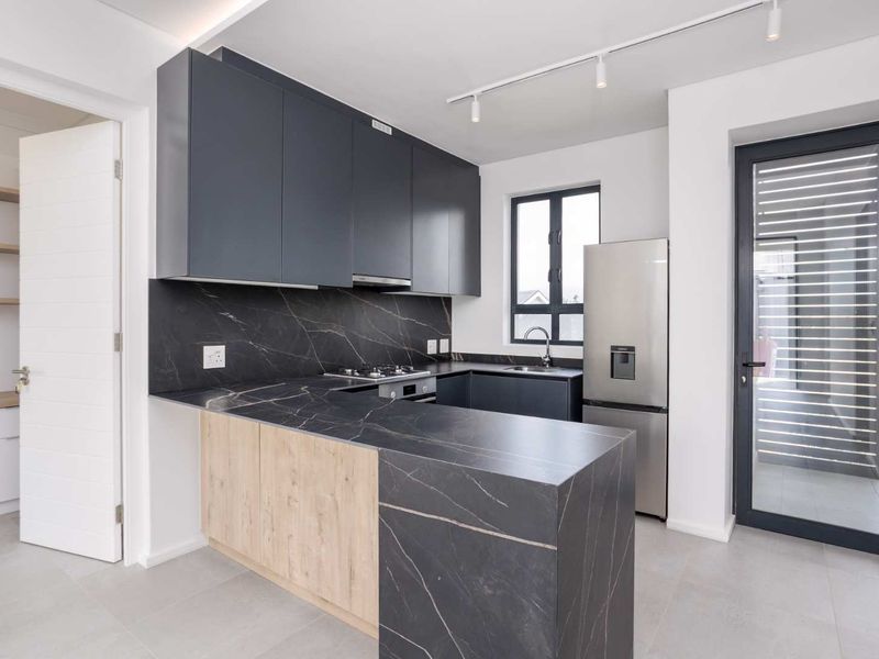 Modern kitchen featuring dark cabinetry, stone countertops, integrated appliances, and an adjacent pantry, finished with contemporary track lighting and neutral floor tiles.