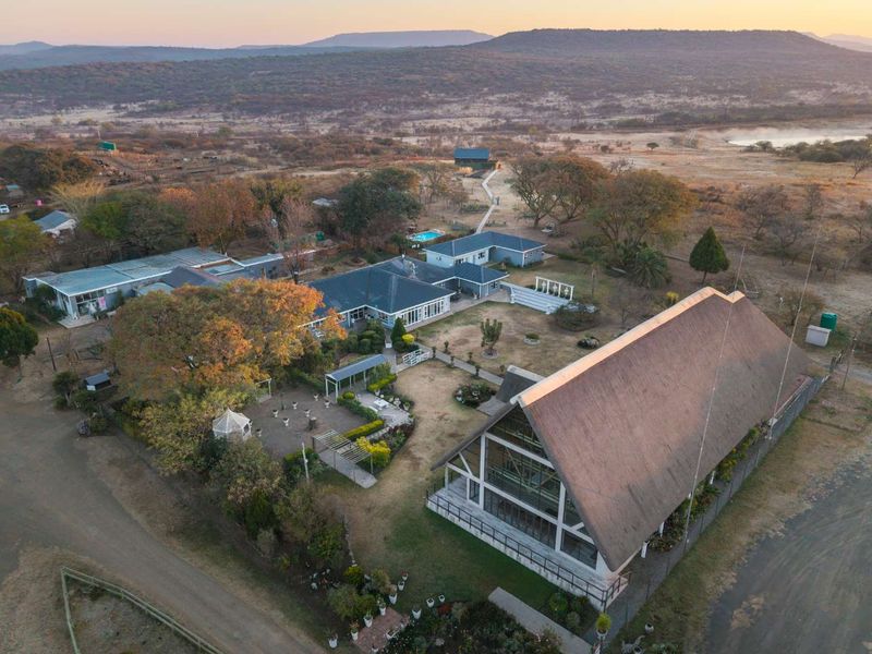 Aerial view of a sprawling guest house property featuring a large thatched-roof building, landscaped gardens, swimming pool, and multiple outbuildings in a rural setting.