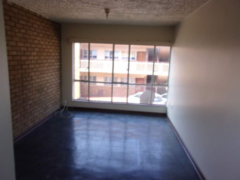 An empty living room featuring an exposed brick accent wall, large window providing natural light, and dark flooring.
