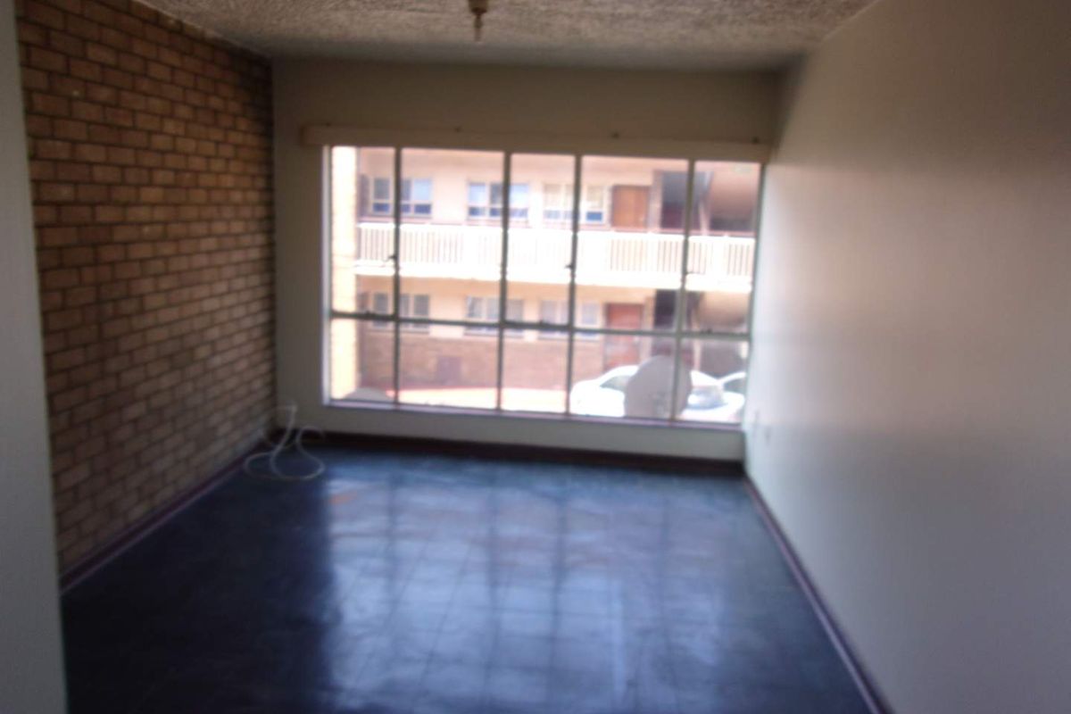 An empty living room featuring an exposed brick accent wall, large window providing natural light, and dark flooring.