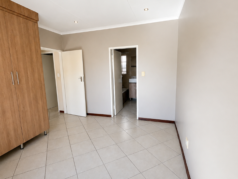 A bright, empty bedroom featuring neutral tiled flooring, built-in wooden wardrobes, and direct access to an en-suite bathroom.