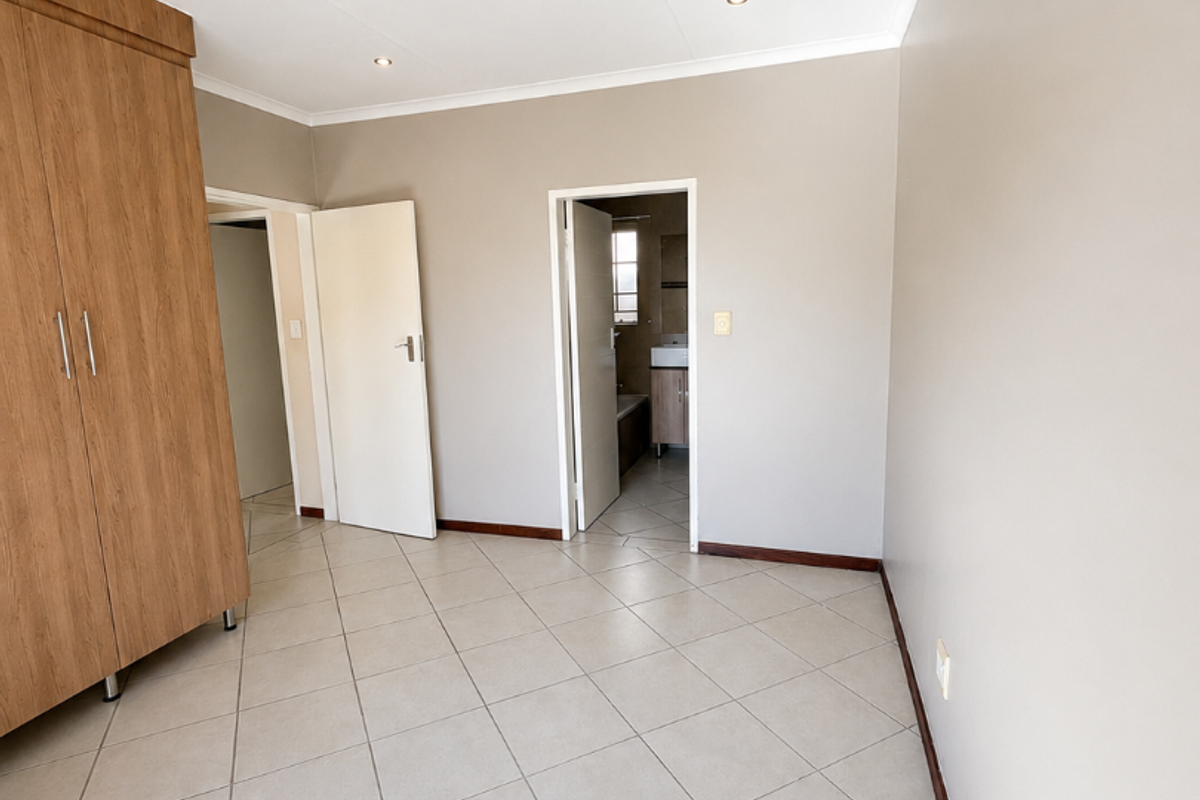 A bright, empty bedroom featuring neutral tiled flooring, built-in wooden wardrobes, and direct access to an en-suite bathroom.