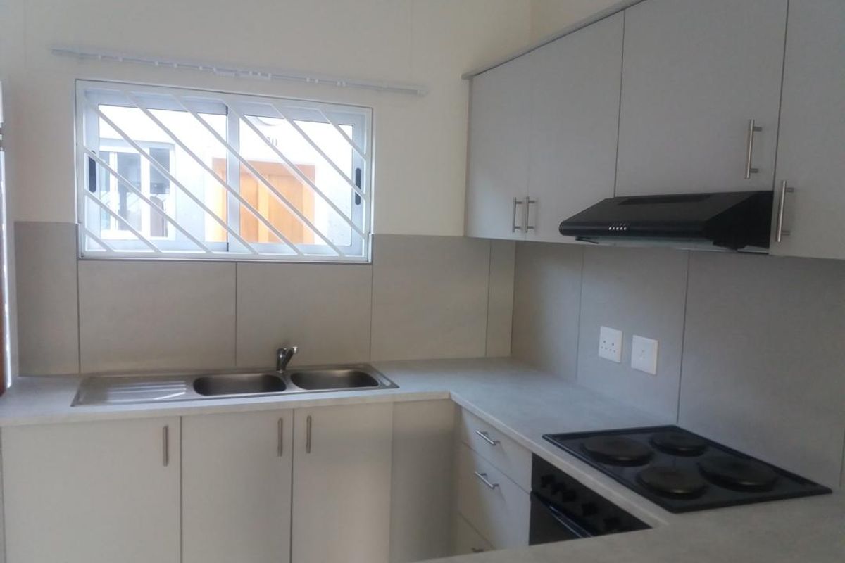 A clean, functional kitchen featuring light grey cabinetry, a built-in electric hob and oven, a double sink, and security bars on the window.