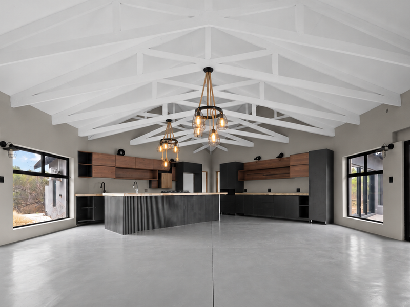 Spacious open-plan kitchen and living area featuring exposed white roof trusses, polished concrete floors, and large sliding doors opening to the surrounding landscape.