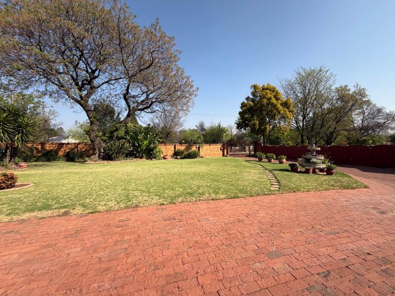 A spacious, well-maintained garden featuring a large lawn, mature trees, a stone fountain, and a brick-paved driveway enclosed by a perimeter wall.