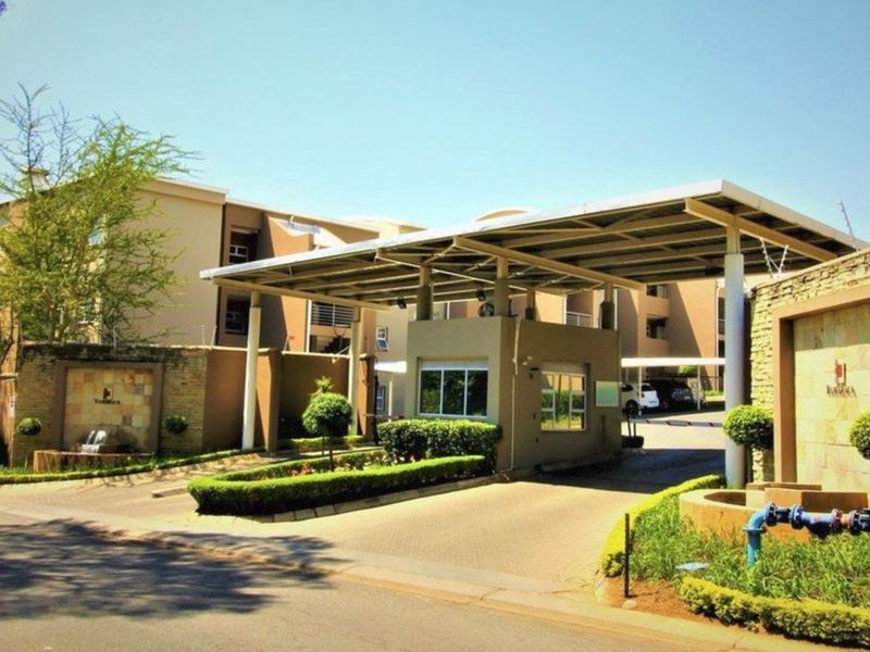 The main entrance to the apartment complex features a secure gatehouse, paved driveway, and well-maintained landscaped gardens with stone-clad architectural details.