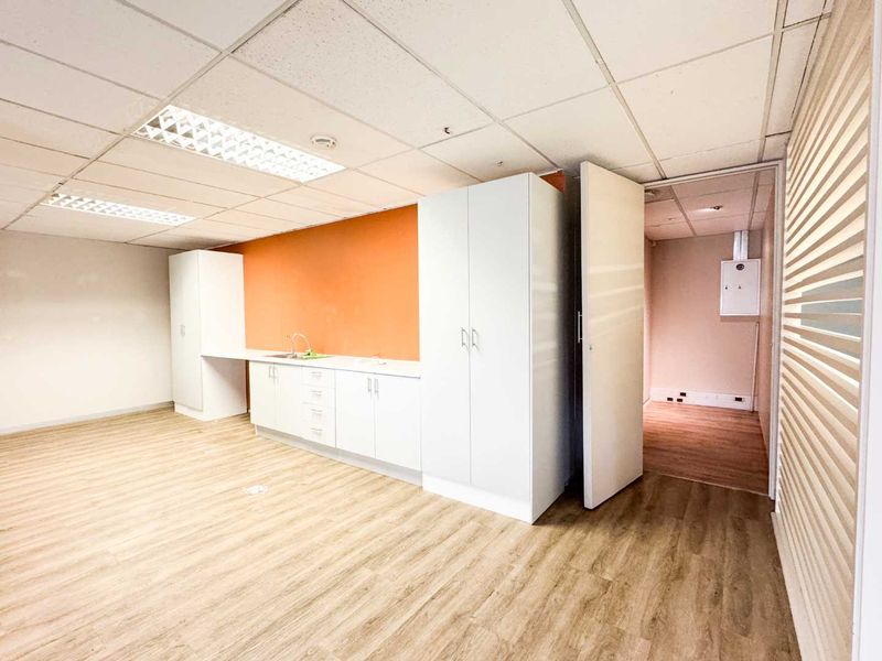 A clean, empty office kitchenette featuring white built-in cabinetry, a stainless steel sink, wood-look laminate flooring, and a drop ceiling with recessed lighting.