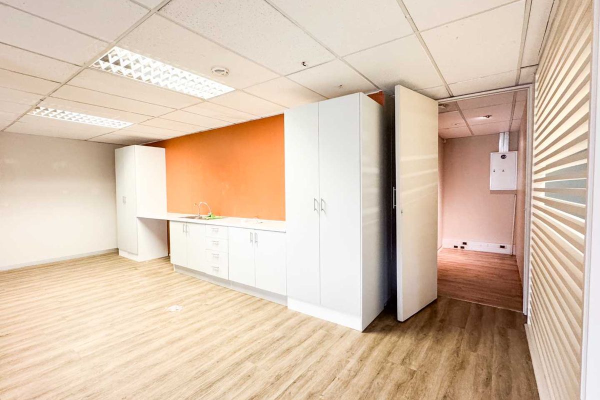 A clean, empty office kitchenette featuring white built-in cabinetry, a stainless steel sink, wood-look laminate flooring, and a drop ceiling with recessed lighting.