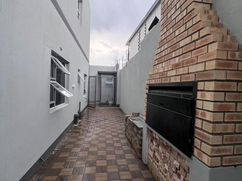 A narrow rear exterior walkway featuring a built-in brick braai, paved flooring, and secure perimeter walling with electric fencing.