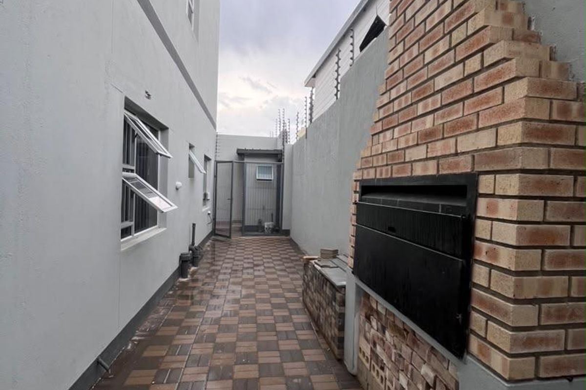A narrow rear exterior walkway featuring a built-in brick braai, paved flooring, and secure perimeter walling with electric fencing.