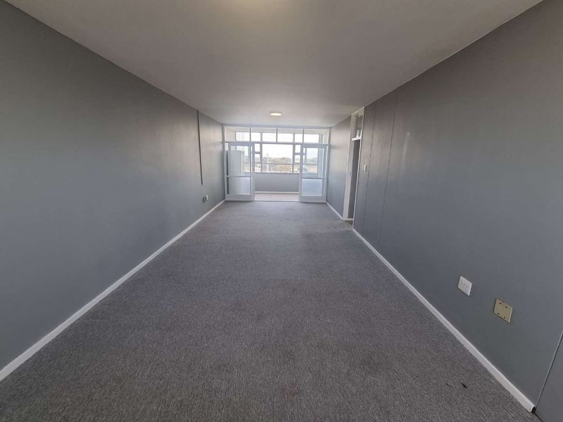 An empty, rectangular living area featuring grey carpeting, neutral walls, and glass-paneled doors leading to an enclosed balcony.
