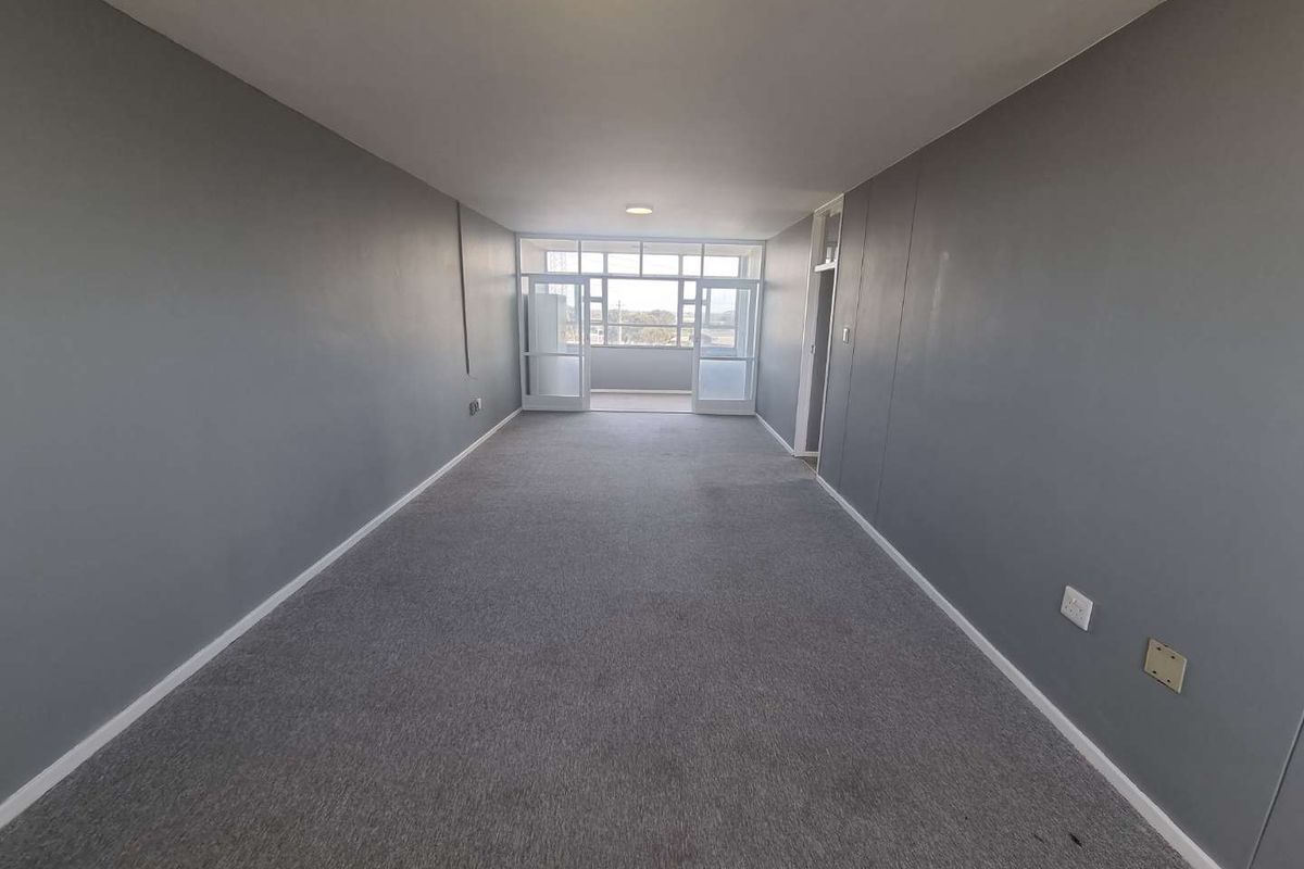 An empty, rectangular living area featuring grey carpeting, neutral walls, and glass-paneled doors leading to an enclosed balcony.